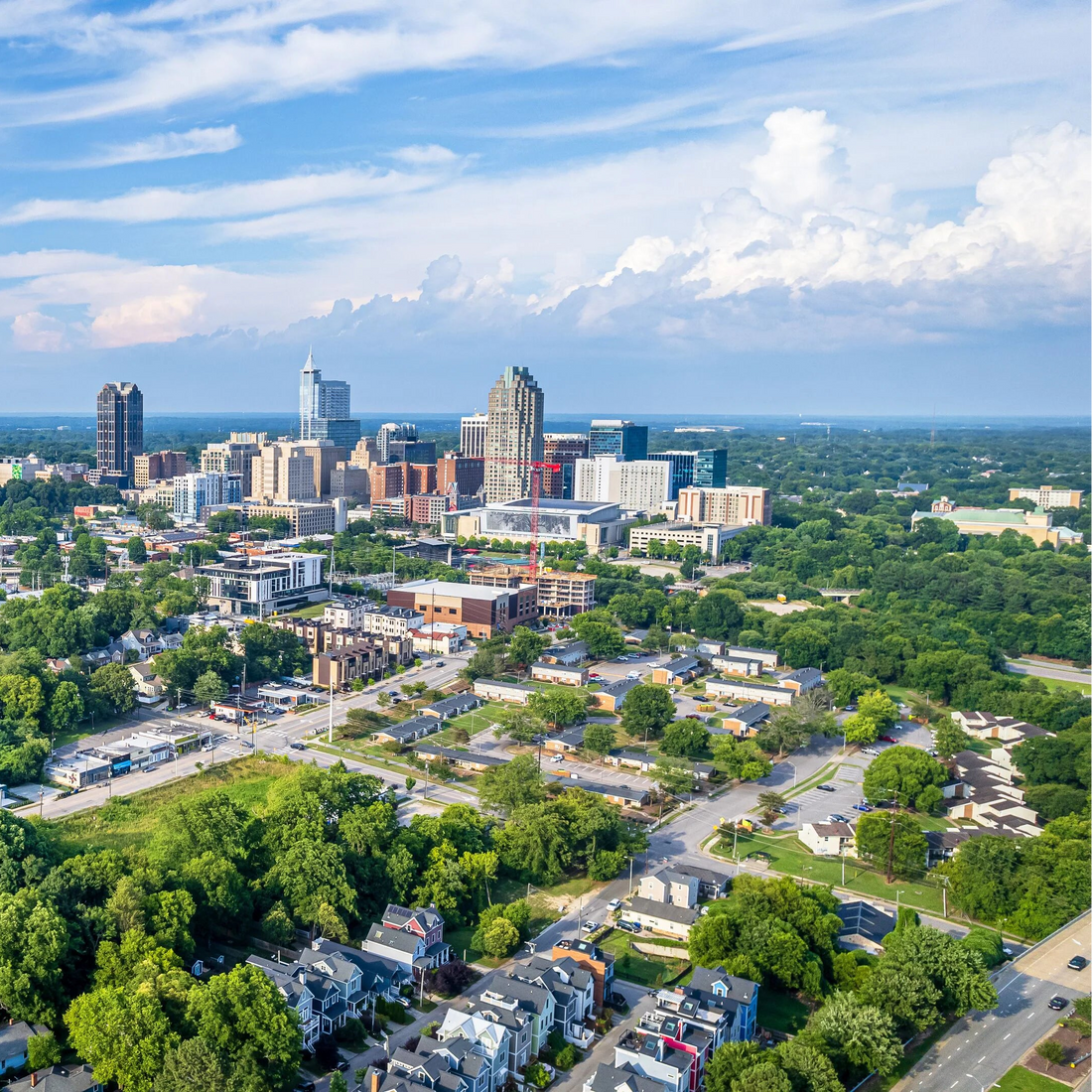 Aerial view of a raleigh, nc with modern buildings and green trees under a blue sky.